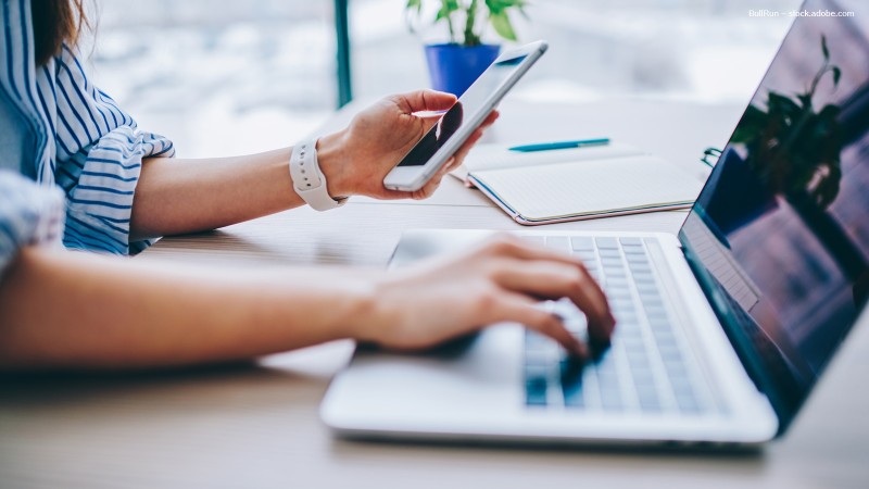 Frau mit Handy in der Hand tippt mit der anderen an einem Laptop.