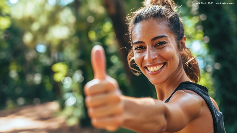 Frau in Sportklamotten ist im Wald und gibt einen Daumen nach oben. 