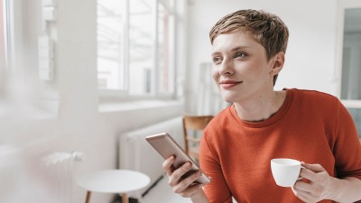 Eine Frau hält in der einen Hand ein Handy und in der anderen eine Tasse.