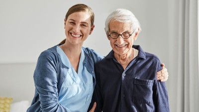 Eine junge Pflegerin hält einen alten Mann im Arm, beide lachen.