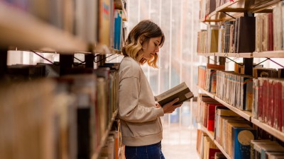 Eine junge Frau steht zwischen Bücherregalen angelehnt und liest ein Buch.