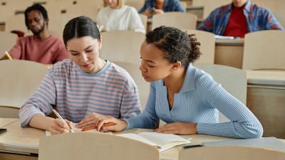 Zwei junge Frauen sitzen in einem Hörsaal und lernen. Hinter ihnen sind weitere Studierende zu sehen.
