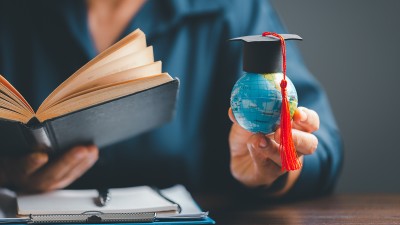 Eine Studierende hält in einer Hand ein Buch, in der anderen einen kleinen Globus mit einer Abschluss-Kappe darauf.
