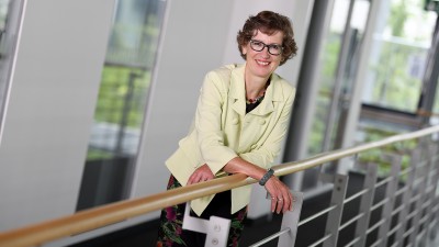 Vorständin Gertrud Prinzing steht an ein Geländer gelehnt, Blick nach vorne, lächelnd, in blassgelbem Blazer