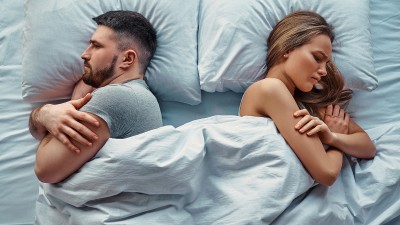 Ein junges Paar liegt im Bett und dreht sich gegenseitig die Rücken zu.