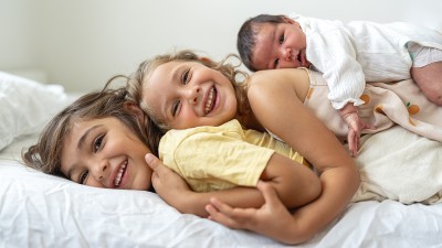 Eine junge Familie – bestehend aus Vater, Mutter und Baby – liegt auf einem weißen Boden, das Baby in der Mitte, die Eltern drum herum und schauen es an.