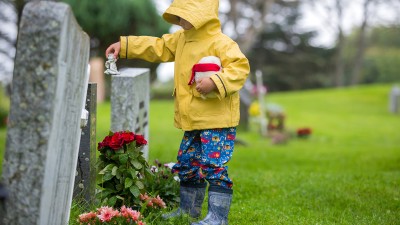 Ein kleiner Junge in einer gelben Regenjacke und blauen Gummistiefeln steht vor einem Grab und stellt eine Engelsfigur darauf ab.