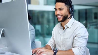 Ein junger Mann in einem Callcenter sitzt an einem Computer und tippt auf der Tastatur. Er trägt ein Headset und lächelt.