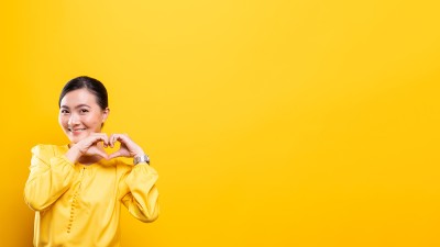 Eine Frau mit gelber Bluse und dunklem Haar steht vor einem gelben Hintergrund. Mit ihren Händen formt sie ein Herz, sie lächelt.