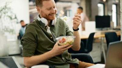 Ein junger Mann mit Kopfhörern um den Hals sitzt an seinem Arbeitsplatz im Büro und isst einen Salat.