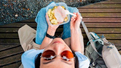 Blick von oben auf eine junge Frau, die auf dem Boden sitzt und isst. Sie trägt eine Sonnenbrille, schaut in die Kamera und hält eine Lunchbox mit Essen in der Hand.