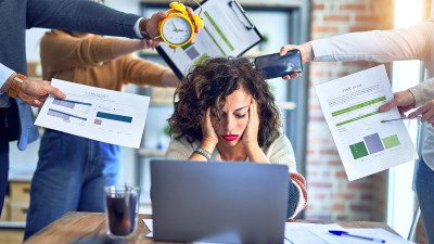 Eine Frau sitz vor ihrem Laptop, den Kopf in den Händen, sie blickt nach unten. Um sie herum stehen Kolleginnen und Kollegen, die ihr verschiedene Dinge wie Klemmbretter mit Unterlagen, einen Wecker oder ein Handy reichen.