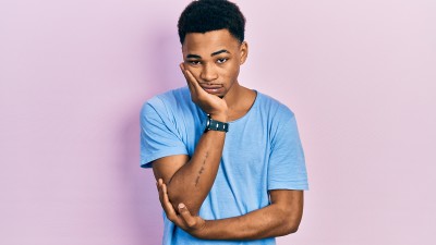 Ein junger Mann in einem hellblauen Shirt hat die Arme vor der Brust verschränkt und stützt mit einer Hand sein Kinn. Er blickt gelangweilt in die Kamera.