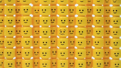 Die gelben Köpfe von Legofiguren übereinander- und nebeneinandergestapelt. Sie lächeln alle – nur der Kopf in der Mitte hat einen traurigen Gesichtsausdruck.