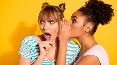 Zwei junge Frauen vor gelbem Hintergrund: Die Frau rechts flüstert der Frau links etwas ins Ohr. Die Frau links hat dabei den Mund geöffnet und wirkt ungläubig.