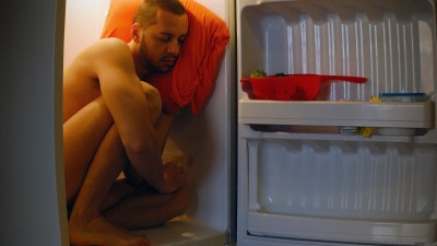 Ein Mann sitzt unbekleidet im Kühlschrank. Sein Kopf lehnt an das Innere der Kühlschranktür auf einem Kissen, seine Augen sind geschlossen.