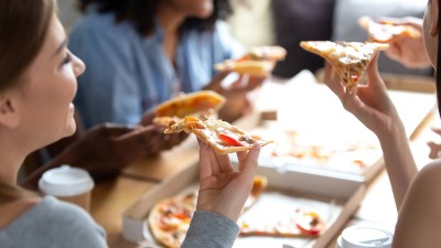 Eine Gruppe von jungen Menschen sitzt gemeinsam am Tisch und genießt Pizza. Im Vordergrund hält eine Person ein Stück Pizza in der Hand, während andere ebenfalls Pizza essen. Getränke in Pappbechern stehen auf dem Tisch.