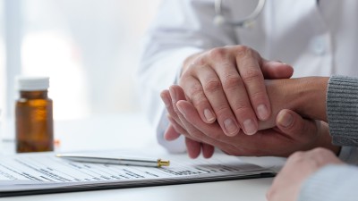 Nahaufnahme von zwei Menschen, die an einem Tisch sitzen: Der Mann hält die Hand einer Frau in seinen Händen. Er trägt einen weißen Arztkittel, sie einen grauen Strickpullover. Vor ihm liegen ein Klemmbrett und ein Kugelschreiber sowie ein Medikamentenfläschen, eine Ampulle und ein Blister mit Tabletten.