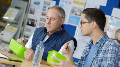 Zwei Männer sitzen gemeinsam an einem Tisch und essen aus grünen Plastikschüsseln. Im Hintergrund sind unscharfe Plakate und eine Pinnwand mit Informationsmaterialien zu sehen.