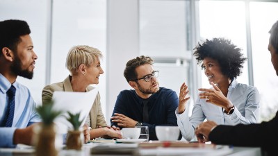 Fünf Geschäftspersonen in einem modernen Konferenzraum, die in einer Besprechung zusammenarbeiten. Zwei Frauen diskutieren, während die anderen Teilnehmer aufmerksam zuhören. Auf dem Tisch liegen Dokumente und ein Laptop.