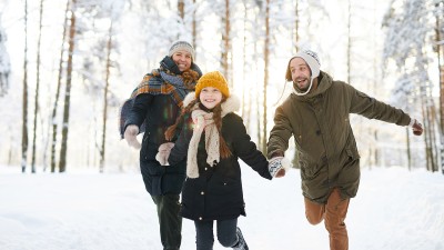 Familie läuft lachend durch einen verschneiten Wald und genießt die Winterlandschaft.