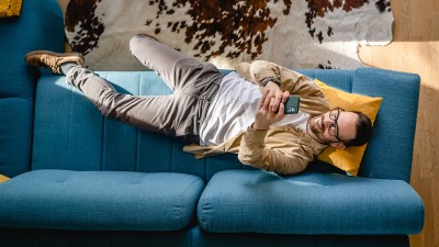 Blick von oben auf einem Mann, der auf einem blauen Sofa liegt. Er hält sein Smartphone in den Händen und schaut auf das Display.