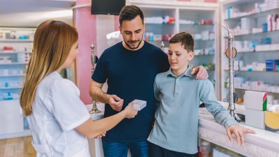 Ein Vater mit seinem Sohn lässt sich in einer Apotheke von einer Apothekerin ein Medikament erklären.