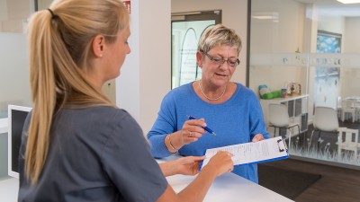 Eine Frau füllt in einer Arztpraxis mit Hilfe einer Angestellten einen Fragebogen aus.