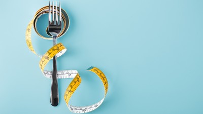 Auf hellblauen Hintergrund liegt eine Gabel, umschlungen von einem Maßband.