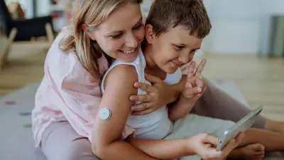 Eine Mutter und ihr Sohn mit einem Glucosesensor lächeln in eine Handykamera.