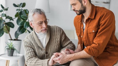 Älterer Mann sitzt auf einem Sofa, ein jüngerer Mann hält ihm die Hand.