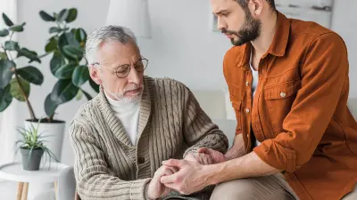 Älterer Mann sitzt auf einem Sofa, ein jüngerer Mann hält ihm die Hand.