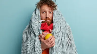 Ein Mann steht in eine Decke gewickelt vor einem hellblauen Hintergrund. Er hat ein Fieberthermometer im Mund und hält in den Händen eine Wärmflasche und eine Zitrone.
