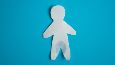 Eine weiße Papierfigur auf hellblauem Hintergrund mit einem nassen Fleck im Schritt