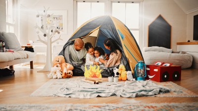 Eine Familie sitzt in einem Indoor-Zelt: Die Eltern rechts und links von den zwei Töchtern, vor ihnen Spielsachen und ein Lagerfeuer aus Bastelpapier.