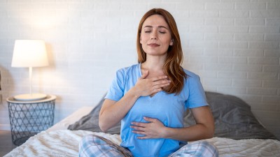 Eine Frau sitzt auf dem Bett, atmet tief, hält eine Hand auf die Brust, die andere auf den Bauch und sieht entspannt aus.