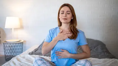 Eine Frau sitzt auf dem Bett, atmet tief, hält eine Hand auf die Brust, die andere auf den Bauch und sieht entspannt aus.