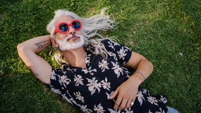 Ein älterer, lässiger Mann mit langen grauen Haaren, roter Sonnenbrille und Hawaiihemd liegt auf einer Wiese und entspannt sich.