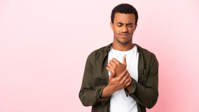 Ein junger Mann steht vor einem rosafarbenen Hintergrund und hält sich mit schmerzverzerrtem Gesicht die Hand
