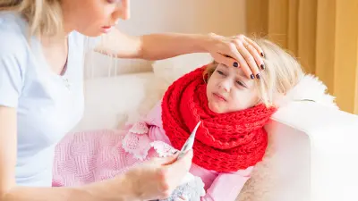 Ein krankes Mädchen liegt auf dem Sofa, die Mutter fühlt die Stirn und hält ein Fieberthermometer.