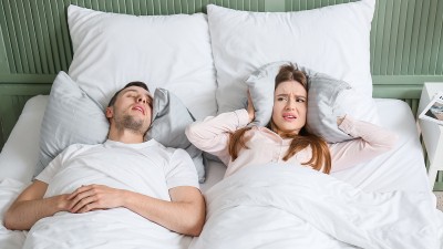 Ein junges Paar liegt im Bett. Der Mann schnarcht, die Frau ist wach und drückt sich ein Kissen auf die Ohren.