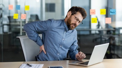 Ein Mann am Computer sitzend hält sich vor Schmerz den unteren Rücken.