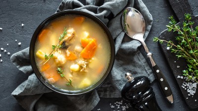 Blick von oben auf eine Suppe mit Gemüseeinlage, daneben liegen ein Löffel, eine Pfeffermühle und frische Kräuter.